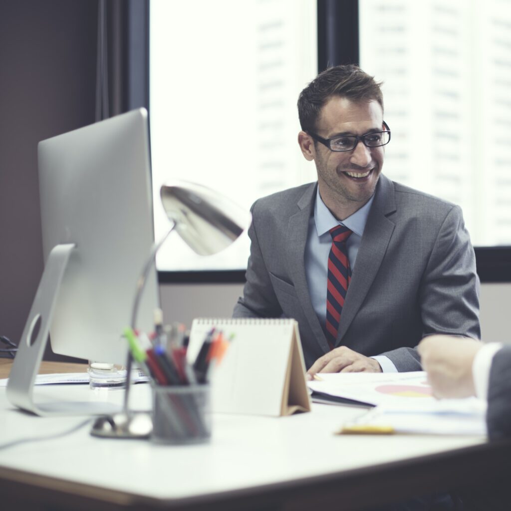 A businessperson sits at their desk and smiles at someone off camera.